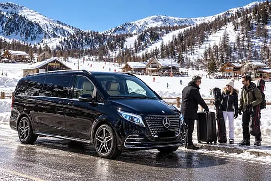 Un van noir et des voyageurs dans une station de ski enneigée, au pied de montagnes.
